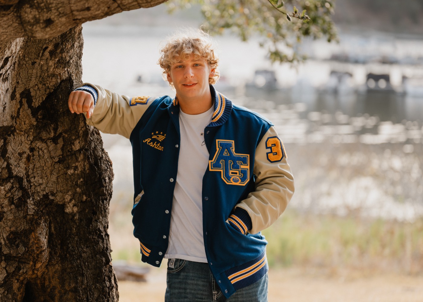 Senior portrait in a varsity letterman jacket under an oak tree by the lake — Central Coast senior photographer
