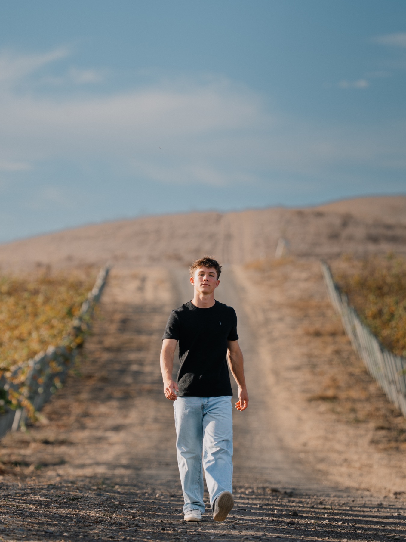 Senior walking toward the camera down a vineyard road under a wide blue sky — Central Coast senior photographer